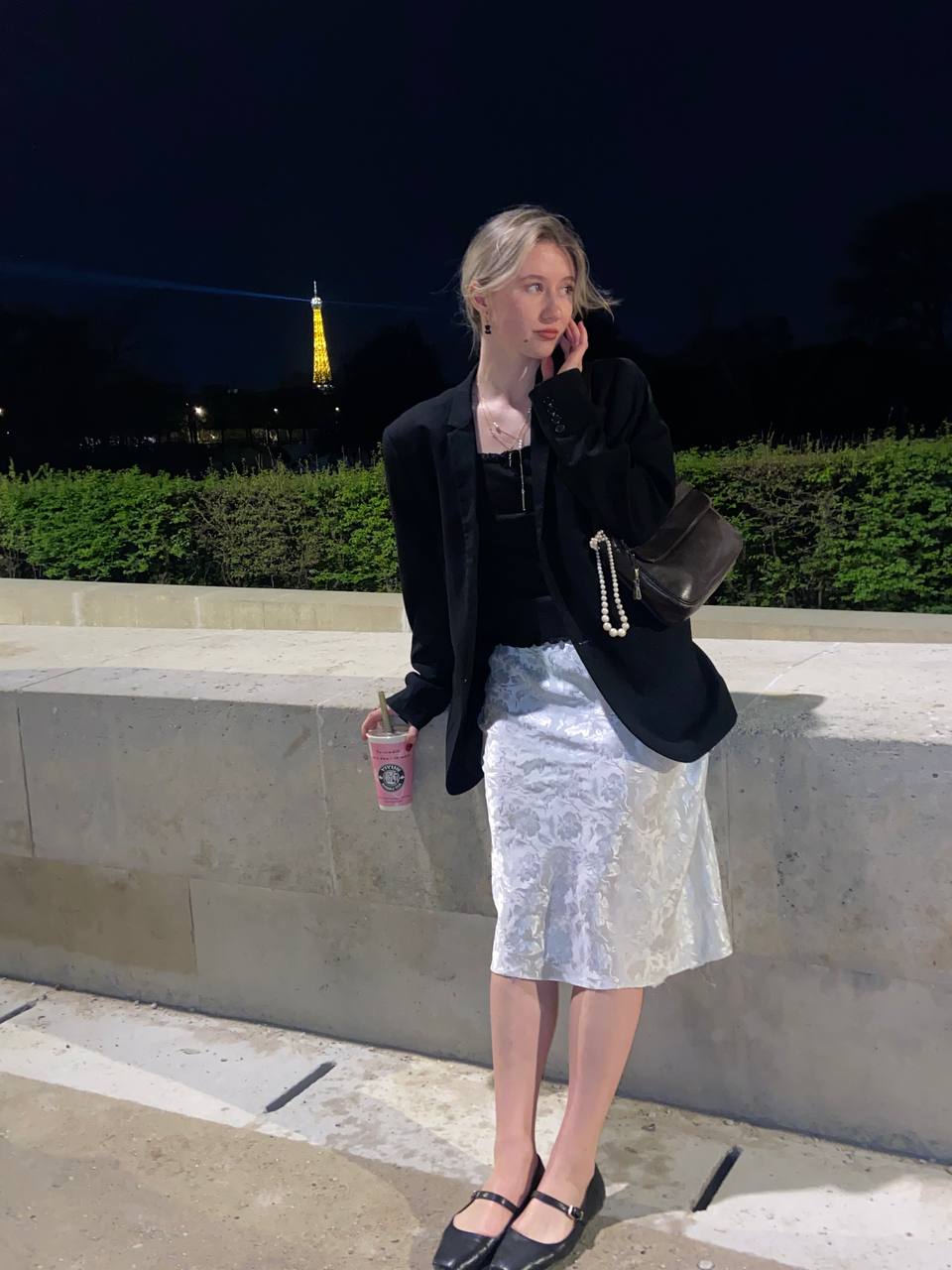 Девушка стоит на фоне Эйфелевой башни в белой юбке и черном пиджаке. Из аксессуаров – черная кожаная сумка с жемчужной подвеской.
